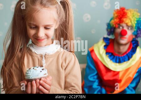 Une petite fille souriante tient le gâteau à la main et fait un souhait. Clown drôle sur le fond. Banque D'Images