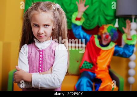 Une petite fille contrariée debout dans la salle de jeux. Clown drôle sur le fond. Banque D'Images