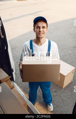 Le livreur en uniforme décharge la voiture avec des colis, service de livraison. Homme debout sur les emballages en carton dans le véhicule, livreur masculin, messager ou picot de transport Banque D'Images