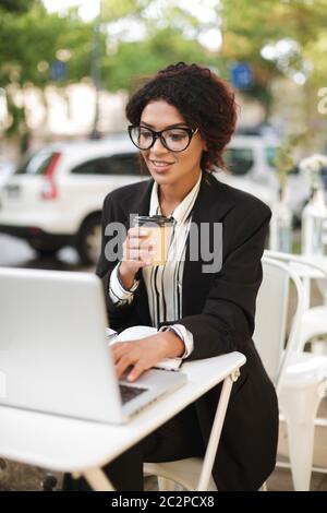 Portrait d'une fille afro-américaine souriante en lunettes, assise à la table du café et travaillant sur son ordinateur portable. Belle dame avec une boisson à cheveux en courbure sombre Banque D'Images