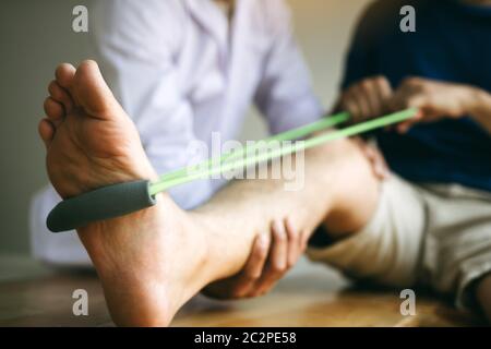 Le patient utilise la bande de résistance qui s'étend sur sa jambe avec un physiothérapeute aide dans la salle de clinique. Banque D'Images