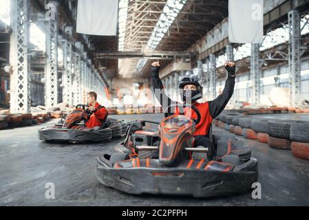 Kart coureur en casque levé ses mains, gagnant, vue avant, karting auto sport en intérieur. Course de vitesse sur une piste de karting étroite avec barrière pour pneus. Rapide Banque D'Images