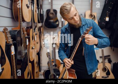Jeune barbu qui choisit la guitare acoustique dans un magasin de musique. Assortiment dans la boutique d'instruments de musique, musicien masculin Banque D'Images