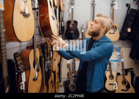 Jeune barbu qui choisit la guitare acoustique dans un magasin de musique. Assortiment dans la boutique d'instruments de musique, musicien masculin Banque D'Images