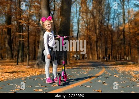 Adorable adorable adorable petite école caucasienne portrait de fille ayant plaisir à faire du vélo de scooter rose de kick dans le parc de ville d'automne or ou la forêt à l'extérieur. Heureux Banque D'Images