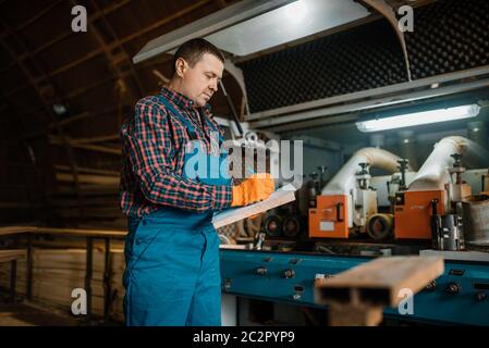 Le menuisier en uniforme tient un cahier, une machine à bois sur fond, l'industrie du bois, la menuiserie. Traitement du bois en usine, sciage de forêt en bûcherons Banque D'Images