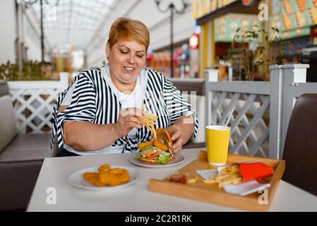 Grosse femme manger des calories des aliments dans le restaurant du centre commercial. L'excès de personne de sexe féminin à la table avec l'ordure dîner, problème d'obésité Banque D'Images