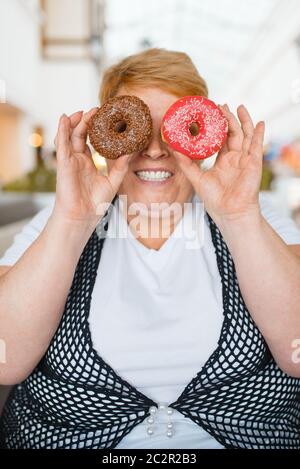 Femme grasse tenant des beignets au lieu des yeux dans le restaurant du centre commercial, nourriture malsaine. Femme en surpoids à la table avec dîner de pourriels, problème d'obésité Banque D'Images