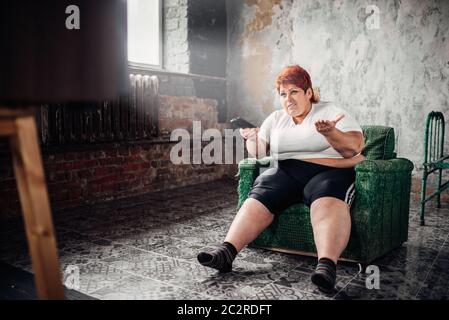 Grosse femme est assise dans un fauteuil et regarde la télévision. Mode de vie malsain, l'obésité Banque D'Images