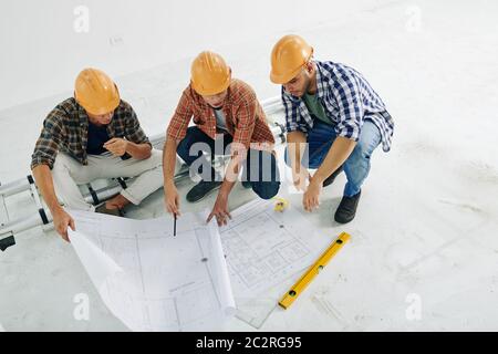 Photo horizontale en grand angle des ouvriers de la construction moderne assis sur des hunkers regardant dans les papiers de plan de construction, l'espace de copie Banque D'Images