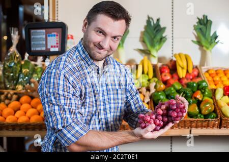 Portrait d'un homme heureux en chemise montrant un bouquet de raisins dans un supermarché. Boîtes avec fruits et légumes sur le fond. Banque D'Images