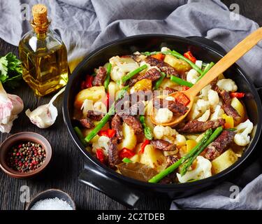 Lanières de boeuf ragoût de pommes de terre neuves, asperges et chou-fleur dans un four hollandais noir sur une table en bois, vue sur le paysage Banque D'Images