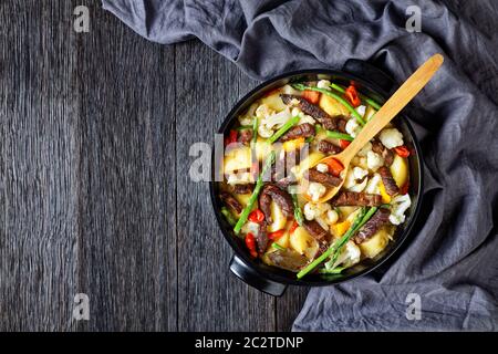 Lanières de bœuf épicées ragoût de pommes de terre neuves, asperges et chou-fleur dans un four hollandais noir sur une table en bois, cuisine américaine, vue sur le paysage, plat la Banque D'Images