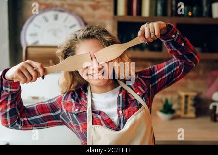 Femme au foyer ludique en tablier couvre ses yeux avec spatules en bois, cuisine intérieur sur arrière-plan. Heureux femme cuisinière faisant des choix de nourriture végétarienne, Banque D'Images