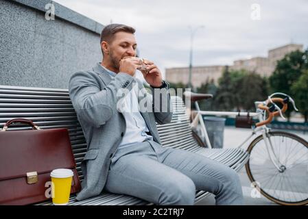 Homme d'affaires avec vélo mange déjeuner sur le banc dans le bâtiment de bureau dans le centre-ville. Personne d'affaires en transport écologique dans la rue de la ville, style urbain Banque D'Images