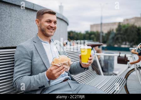 Homme d'affaires avec vélo mange déjeuner sur le banc à l'immeuble de bureaux dans le centre-ville. Personne d'affaires en transport écologique dans la rue de la ville, style urbain Banque D'Images