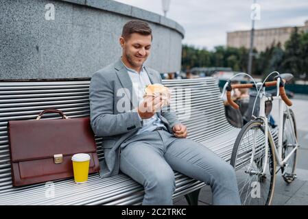 Homme d'affaires avec vélo mange déjeuner sur le banc dans le bâtiment de bureau dans le centre-ville. Personne d'affaires en transport écologique dans la rue de la ville, style urbain Banque D'Images