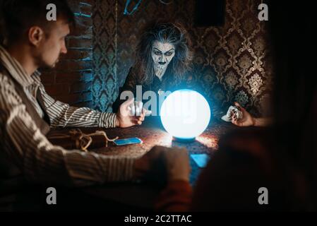 Sorcière effrayante lit un charme magique sur une boule de cristal, jeune homme et femme sur la seance spirituelle. Banque D'Images