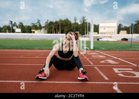 Jogging féminin fatigué assis sur le sol, entraînement sur le stade. Femme qui fait de l'exercice d'étirement avant de courir sur une arène extérieure Banque D'Images