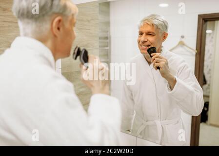 Cheerful woman in bathrobe rase au miroir dans la salle de bains. Mâle mature aux cheveux gris avec personne - Rasoir électrique Banque D'Images