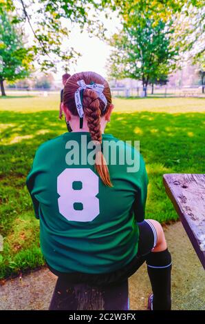 Une jeune fille aux cheveux tressés et portant le maillot de sport/maillot de sport numéro 8 reposant sur un banc de parc après une pratique de football. Banque D'Images