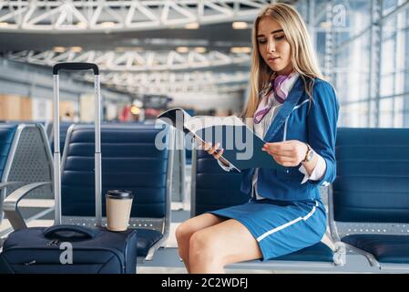 Hôtesse de l'air avec valise assis sur son fauteuil dans la salle d'attente à l'aéroport et se lit magazine. Hôtesse de l'air, avec des bagages de cabine avec un bagage à main, Banque D'Images