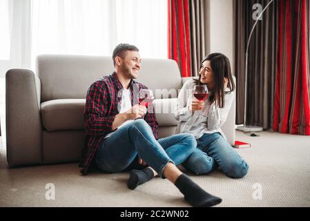 Love couple assis sur le sol contre la table, montres de film et de boissons vin rouge de grands verres, fenêtre et un salon intérieur sur l'arrière-plan. Rom Banque D'Images