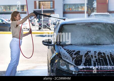 Jeune femme caucasienne conducteur se lavant au lavage manuel de voiture, le nettoyage avec de la mousse, l'eau sous pression. Lavage de voiture en libre-service avec tuyau haute pression Banque D'Images