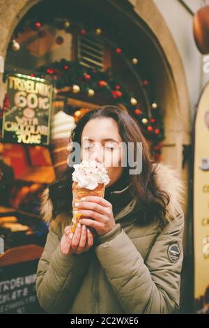 Une belle fille dans une veste chaude mange trdelnik ou Trdlo avec de la crème dans ses mains, en hiver en République tchèque, Prague à Banque D'Images