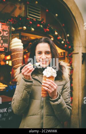 Une belle fille dans une veste chaude mange trdelnik ou Trdlo avec de la crème dans ses mains, en hiver en République tchèque, Prague à Banque D'Images