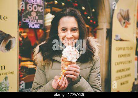 Une belle fille dans une veste chaude mange trdelnik ou Trdlo avec de la crème dans ses mains, en hiver en République tchèque, Prague à Banque D'Images