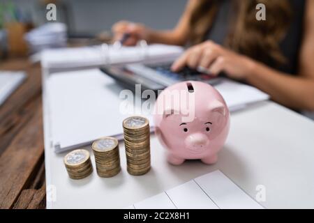 Femme D'Affaires Utilisant Calculatrice Avec Piggybank Et Pile De Pièces Sur Le Bureau Banque D'Images
