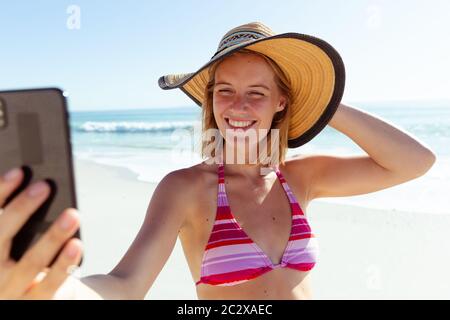 Femme caucasienne à la plage portant un chapeau et prenant selfie avec son smartphone Banque D'Images