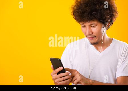 Joyeux homme asiatique aux cheveux raillés, il souriait en écoutant de la musique sur des écouteurs à l'aide d'un smartphone mobile isoler sur fond jaune Banque D'Images