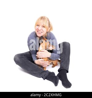 Femme blonde assise sur le sol avec son petit chien brun isolé sur fond blanc dans le studio. Petite race mixte de cric Banque D'Images