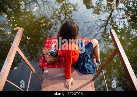 Une jeune femme assise avec son dos sur un pont près d'un lac sur une journée ensoleillée. Plaid, appareil photo, suitcase Banque D'Images