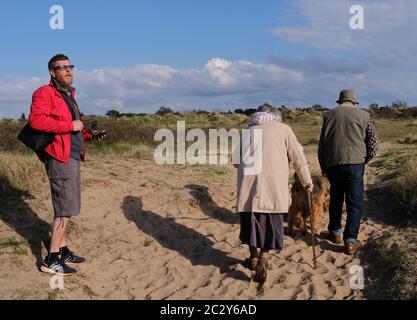 vieux couple avec chien marcher devant un jeune homme en veste rouge, en regardant la mer sur le sentier de la plage, au soleil éclatant avec ciel bleu et de longues ombres Banque D'Images