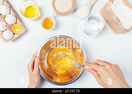 Vue de dessus des mains de femmes fouetter des œufs pour faire de la pâte pour du pain maison à côté des ingrédients sur une table de cuisine blanche. Banque D'Images