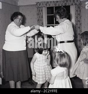 Années 1960, historique, dans une pièce à l'occasion de la fête d'anniversaire d'un enfant, les jeunes enthousiastes s'amusent à jouer à un jeu qui se déroule sous les bras de deux dames, Angleterre, Royaume-Uni. Banque D'Images