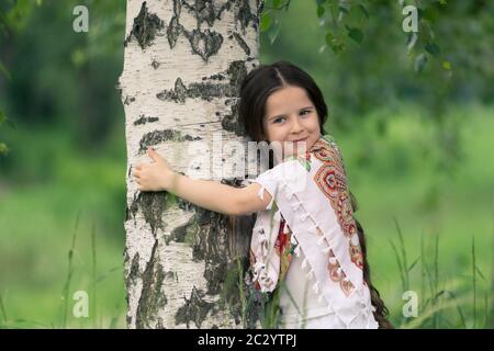 Portrait d'une petite belle fille embrassant un bouleau. Ses épaules sont recouvertes d'un châle aux motifs traditionnels russes. Banque D'Images