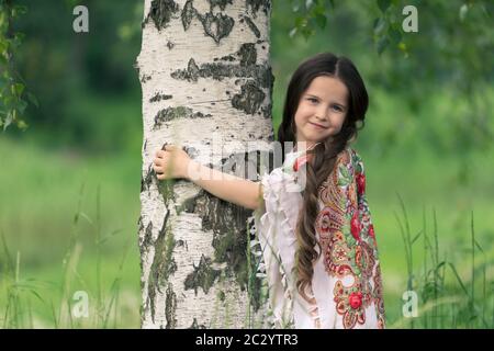 Portrait d'une petite belle fille embrassant un bouleau. Ses épaules sont recouvertes d'un châle aux motifs traditionnels russes. Banque D'Images