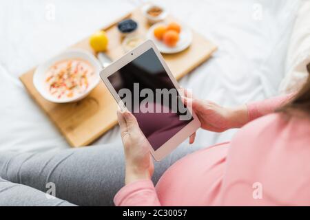 Gros plan de la jeune femme enceinte utilisant un comprimé numérique à côté de flocons d'avoine frais et de fruits sur une petite table au lit. Banque D'Images