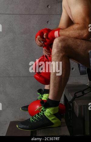 Gros plan sur un boxeur homme mi-adulte fatigué assis après un match d'entraînement, tenant des gants de boxe rouge avec les mains dans des enveloppements protecteurs de poignet, à faible angle Banque D'Images