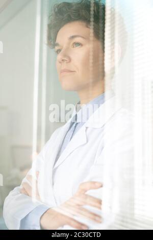 Chercheuse médicale ambitieuse et confiante avec des cheveux courts debout, bras croisés à la fenêtre avec des stores Banque D'Images