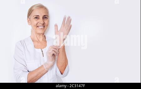 Femme âgée souriante femme mature médecin ou infirmière dans un manteau médical blanc met des gants portant un équipement de protection individuelle isolé. Santé et Banque D'Images