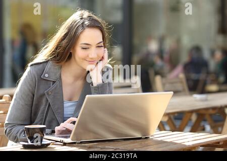 Jeune fille satisfaite regardant le contenu médiatique sur son ordinateur portable sur une terrasse de café une journée ensoleillée Banque D'Images