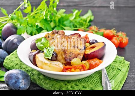 Poulet cuit au four avec tomates, pommes, prunes et raisins dans une assiette sur une serviette vert, ail, persil et basilic sur planche de bois contexte Banque D'Images