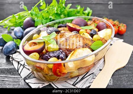 Poulet cuit au four avec tomates, pommes, prunes et raisins dans un torréfacteur de verre sur une serviette, ail, persil et basilic sur fond de planche de bois foncé Banque D'Images