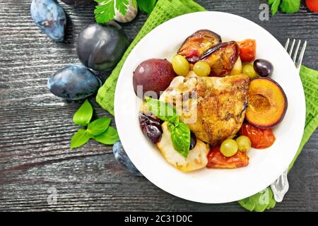 Poulet cuit au four avec tomates, pommes, prunes et raisins dans un plat sur une serviette, ail, persil et basilic sur planche de bois au-dessus de l'arrière-plan Banque D'Images