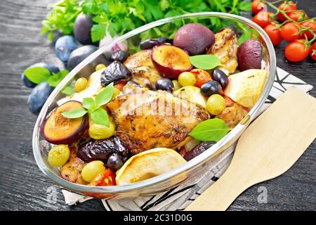 Poulet cuit au four avec tomates, pommes, prunes et raisins dans un torréfacteur de verre sur une serviette, ail, persil et basilic sur fond de planche de bois foncé Banque D'Images